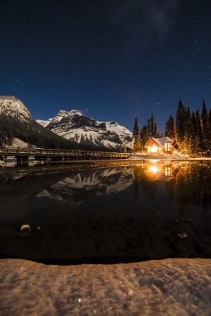 星空を背景に凍るような湖のそばで火に照らされた家が光る写真