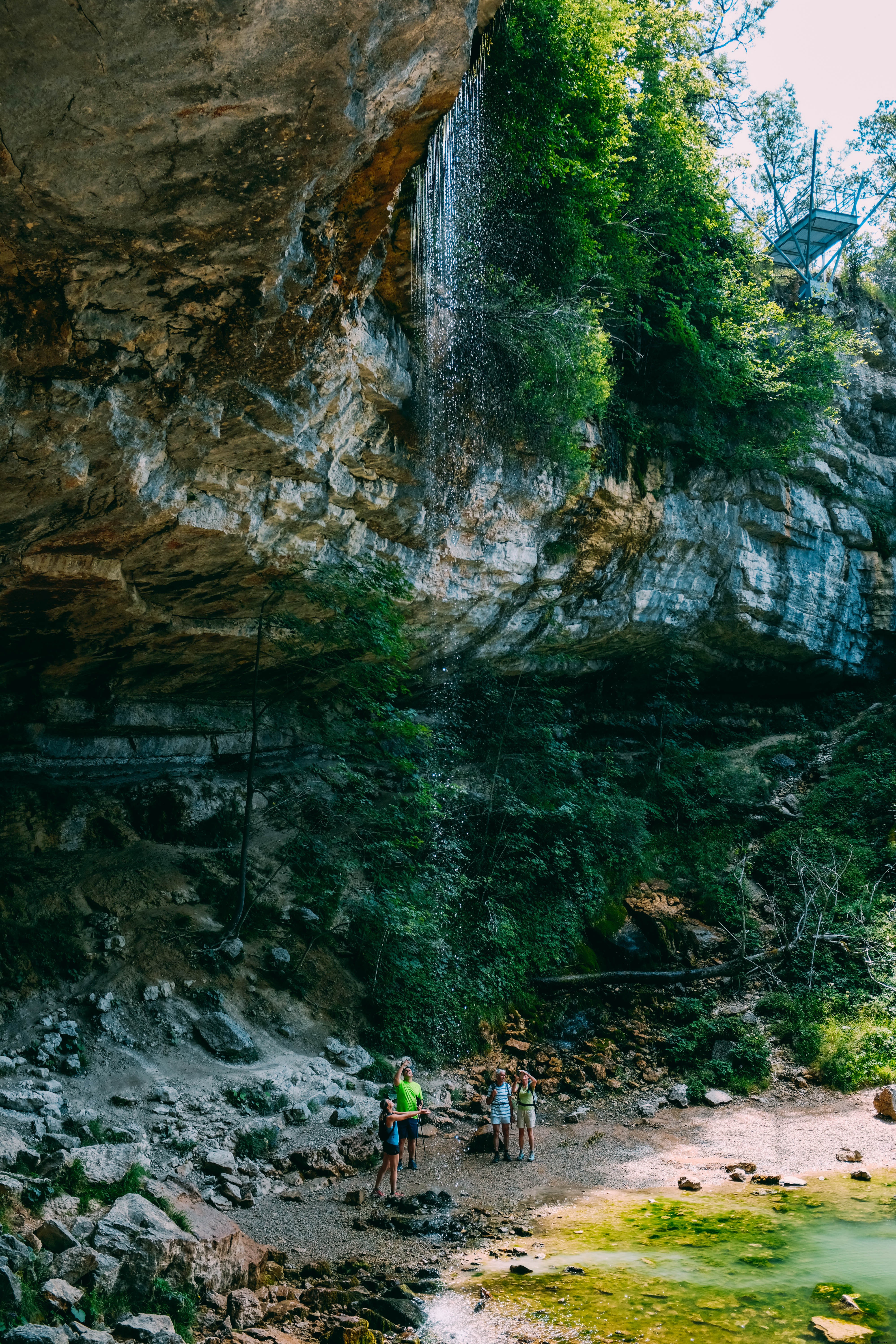 Foto de caminhantes sob um afloramento rochoso e uma pequena cachoeira