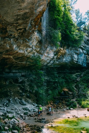 Foto de caminhantes sob um afloramento rochoso e uma pequena cachoeira