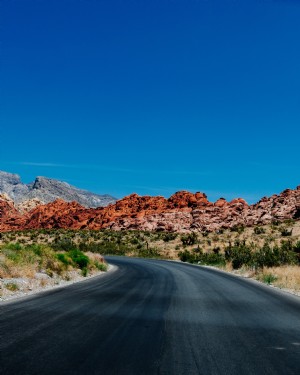 Estrada pavimentada curva com colinas rochosas vermelhas ao fundo. 