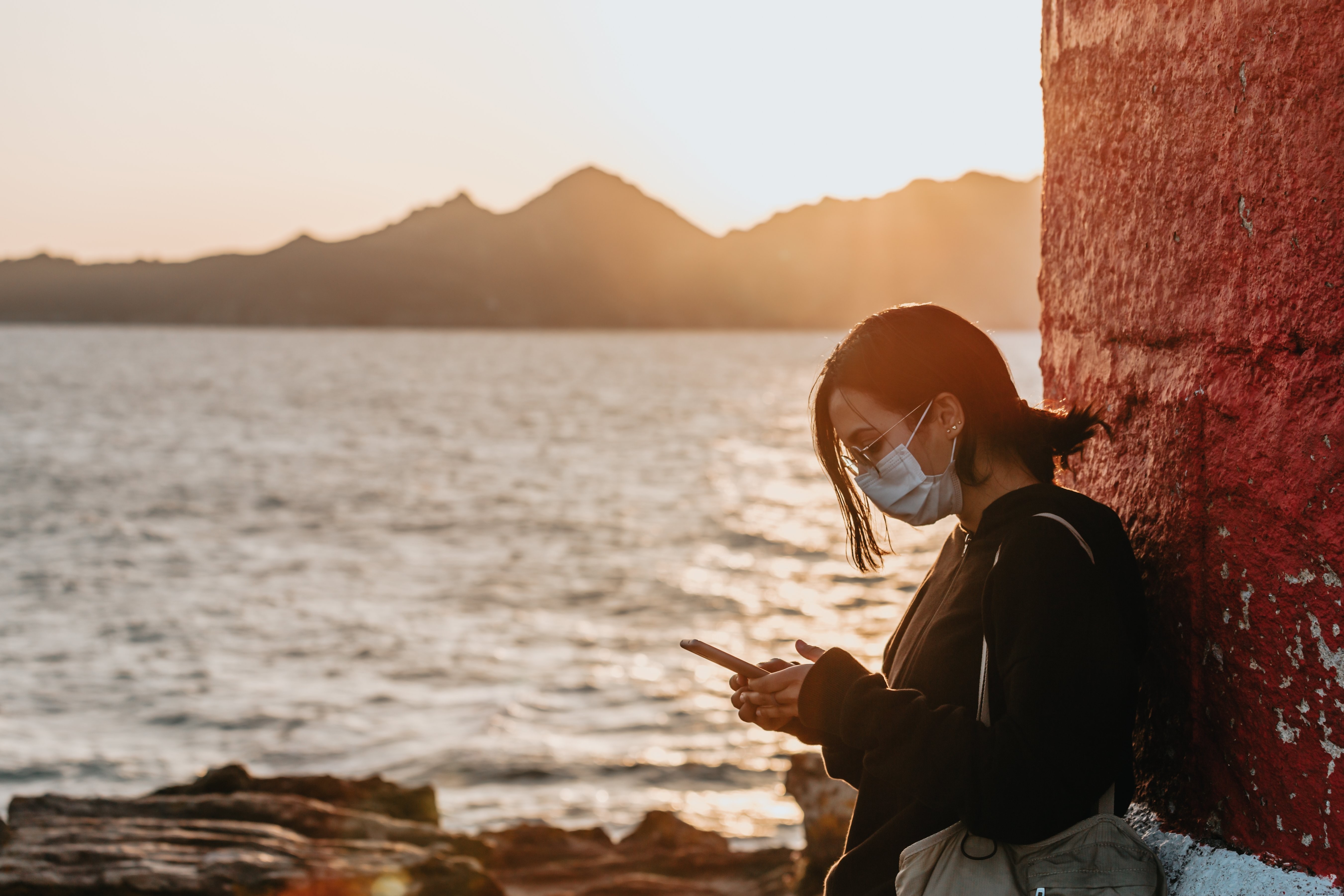 フェイスマスクの女性が水の写真のそばに立っている彼女の携帯電話を見る 