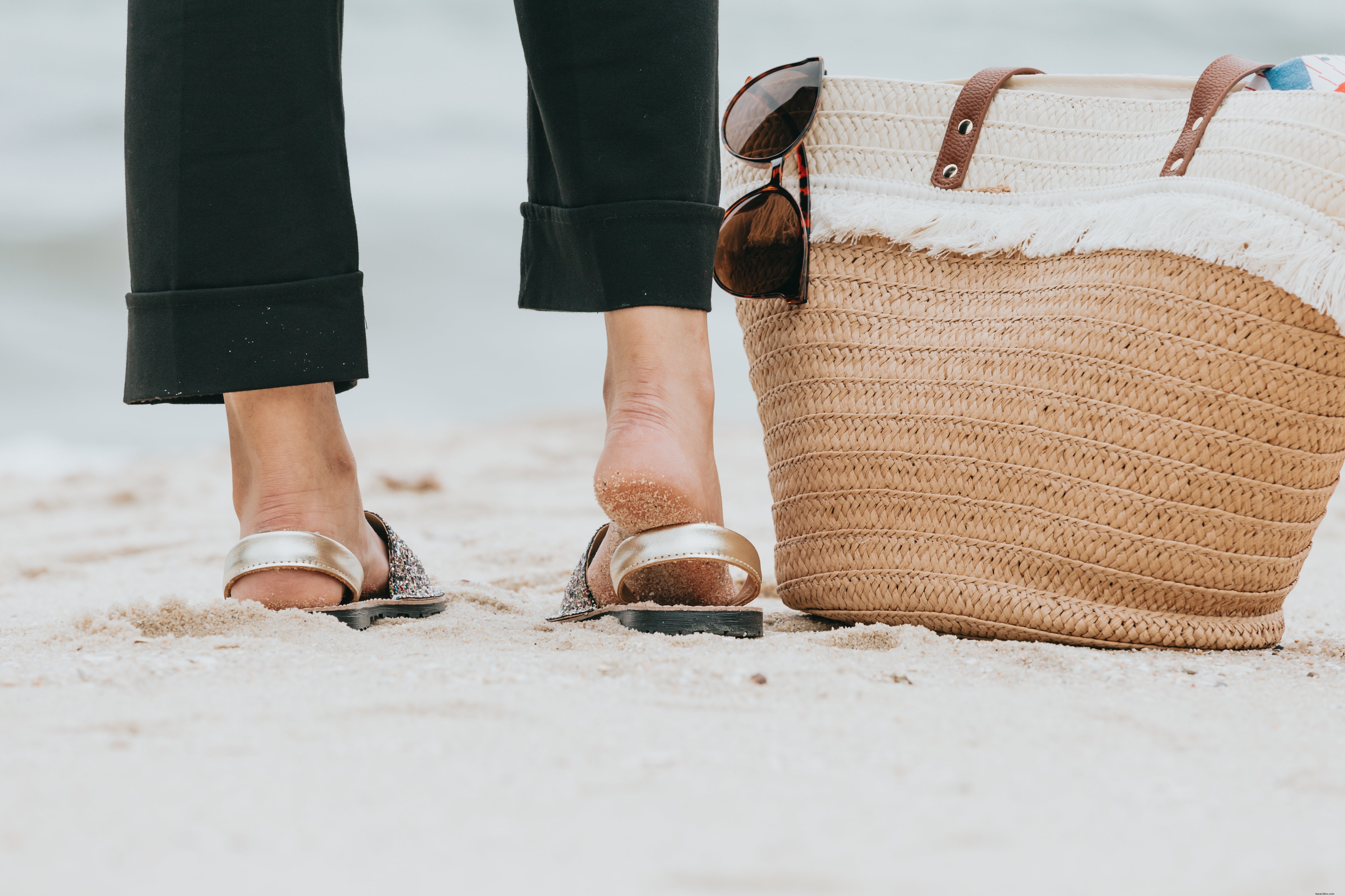 Foto de uma perna em pé em uma praia de areia ao lado de uma sacola de praia. 