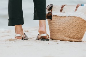 Foto de uma perna em pé em uma praia de areia ao lado de uma sacola de praia. 